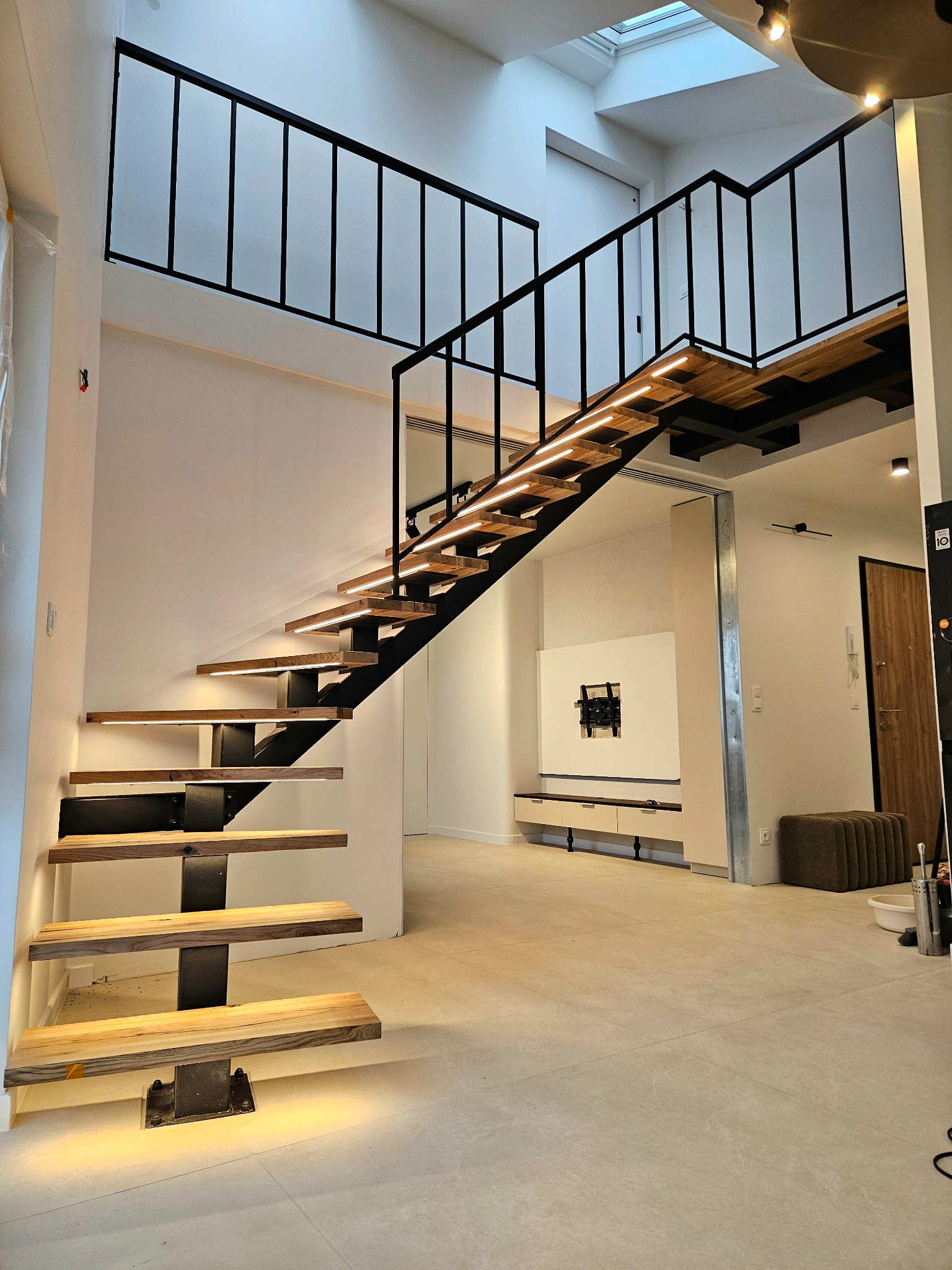 Nowoczesne schody z drewnianymi stopniami i czarną metalową konstrukcją, podświetlane LED, w jasnym wnętrzu z antresolą i oknem dachowym.