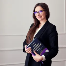 Uśmiechnięta kobieta w okularach i czarnym żakiecie trzyma w rękach fioletową książkę 'Ustawa o gospodarce nieruchomościami - Komentarz' na tle jasnej ściany.