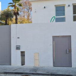 PRO TECH SECURITE SPÓŁKA Z OGRANICZONĄ ODPOWIEDZIALNOŚCIĄ - Nowoczesna, szara brama wjazdowa przesuwna, zintegrowana z białym murem otaczającym posesję, obok szare drzwi zewnętrzne z długim uchwytem, w tle widoczne drzewa i budynki.