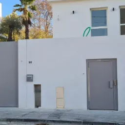 Nowoczesna, szara brama wjazdowa przesuwna, zintegrowana z białym murem otaczającym posesję, obok szare drzwi zewnętrzne z długim uchwytem, w tle widoczne drzewa i budynki.