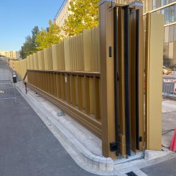 PRO TECH SECURITE SPÓŁKA Z OGRANICZONĄ ODPOWIEDZIALNOŚCIĄ - Złota, przesuwna brama wjazdowa z pionowymi panelami, częściowo otwarta, na tle budynku z licznymi oknami i zielenią drzew.