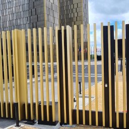 PRO TECH SECURITE SPÓŁKA Z OGRANICZONĄ ODPOWIEDZIALNOŚCIĄ - Nowoczesna brama wjazdowa z pionowych, złotych i czarnych profili, na tle budynku biurowego i flag.