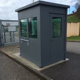 PRO TECH SECURITE SPÓŁKA Z OGRANICZONĄ ODPOWIEDZIALNOŚCIĄ - Szara budka wartownicza z płaskim dachem i dwoma oknami, umieszczona przy bramie wjazdowej na asfaltowym placu.