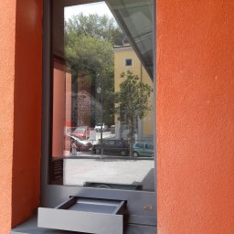 PRO TECH SECURITE SPÓŁKA Z OGRANICZONĄ ODPOWIEDZIALNOŚCIĄ - Okno z podajnikiem, szara rama, pomarańczowa ściana z fakturą, odbicie ulicy w szybie.