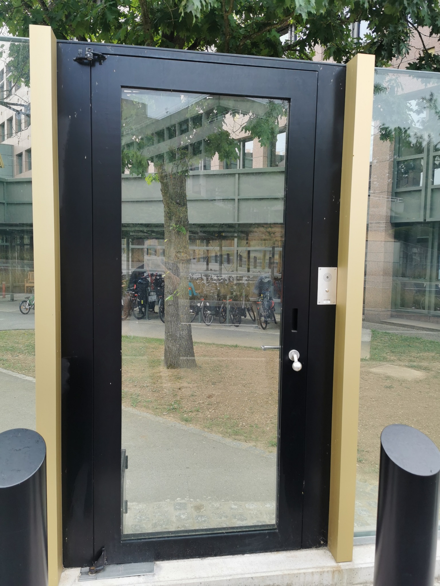 Zewnętrzne przeszklone drzwi w czarnej ramie z beżowymi filarami, widok na parking rowerowy i budynek w tle.