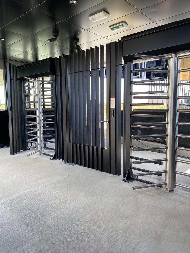 Nowoczesne wejście z obrotowymi barierami metalowymi, czarne elementy konstrukcyjne i srebrne rury, widoczne oświetlenie sufitowe.