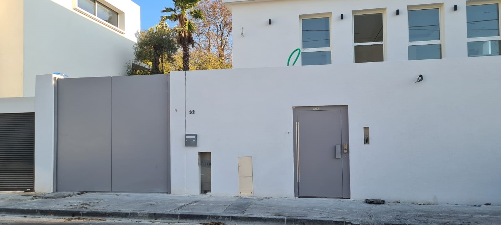 Nowoczesna, szara brama wjazdowa przesuwna, zintegrowana z białym murem otaczającym posesję, obok szare drzwi zewnętrzne z długim uchwytem, w tle widoczne drzewa i budynki.