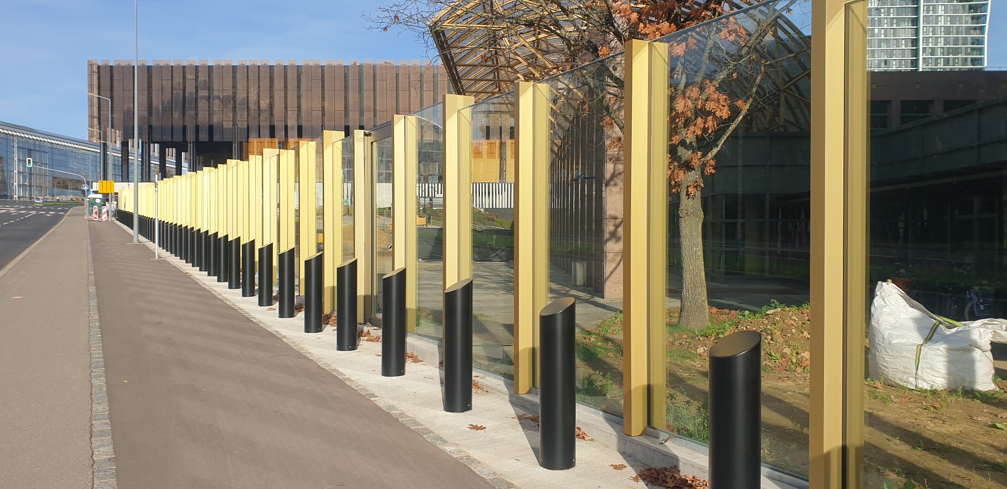 Długa, nowoczesna bariera z pionowych, złotych słupków i przezroczystych paneli, oddzielająca chodnik od jezdni, z budynkami w tle.