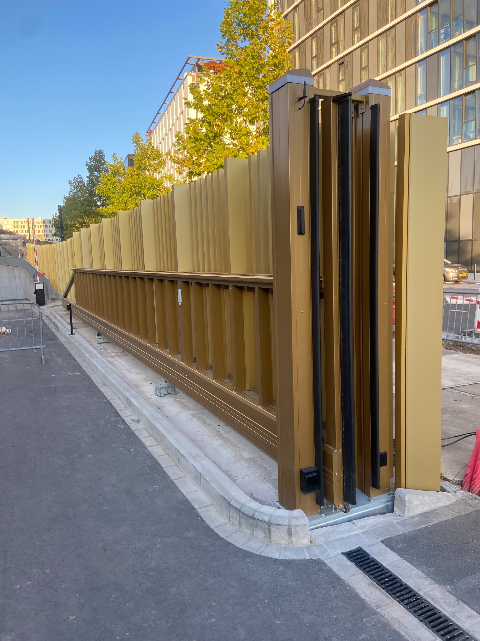 Złota, przesuwna brama wjazdowa z pionowymi panelami, częściowo otwarta, na tle budynku z licznymi oknami i zielenią drzew.