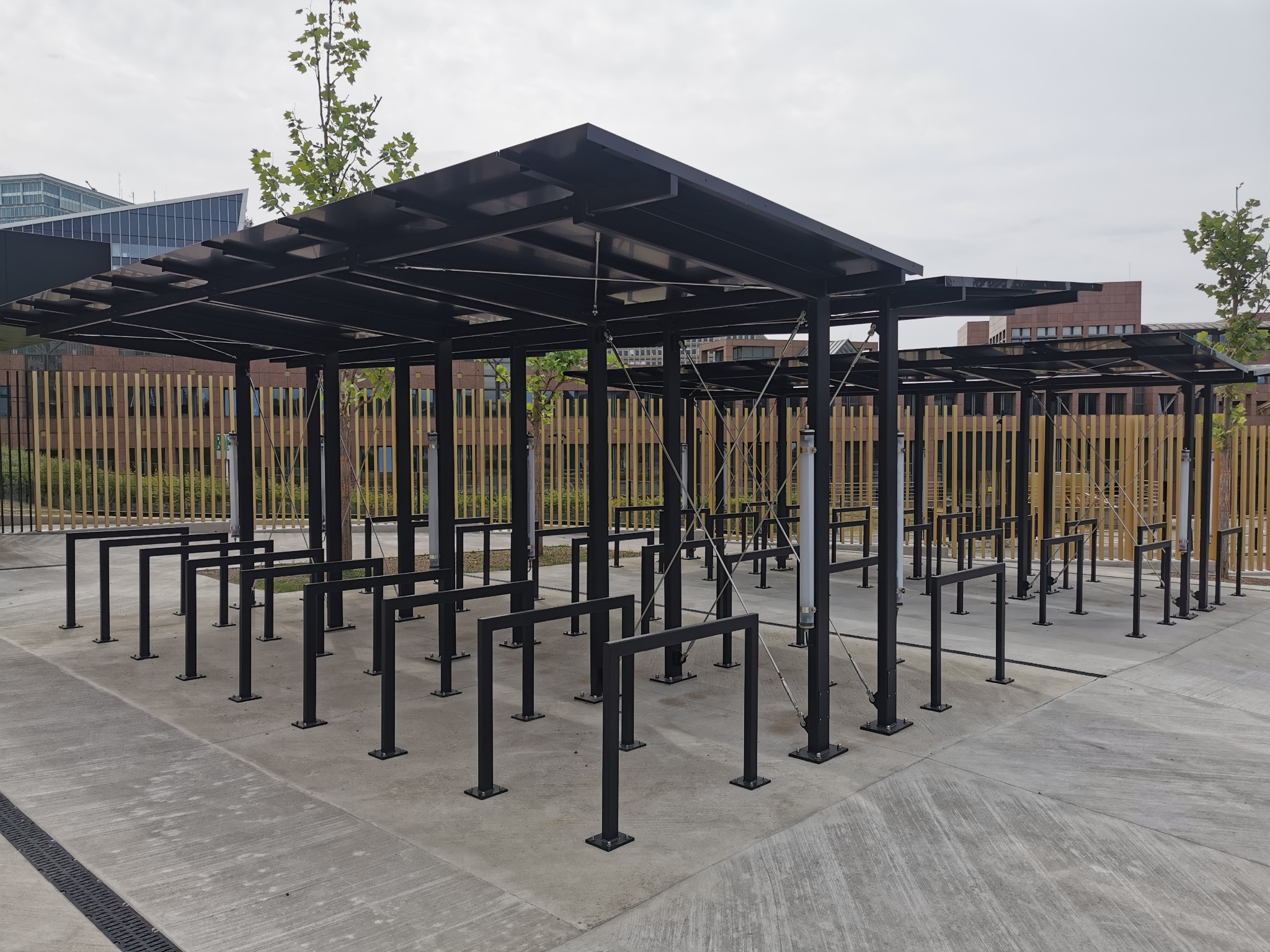 Zadaszony parking rowerowy z metalową konstrukcją, widoczne elementy mocujące i tło z budynkami.