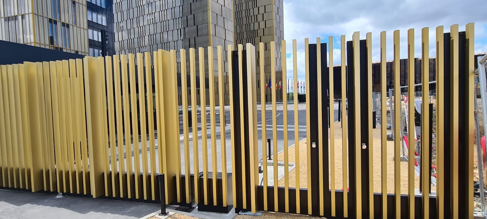 Nowoczesna brama wjazdowa z pionowych, złotych i czarnych profili, na tle budynku biurowego i flag.