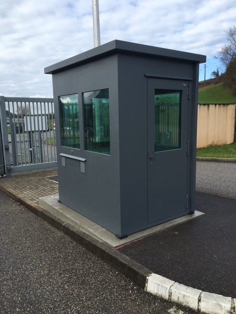 Szara budka wartownicza z płaskim dachem i dwoma oknami, umieszczona przy bramie wjazdowej na asfaltowym placu.