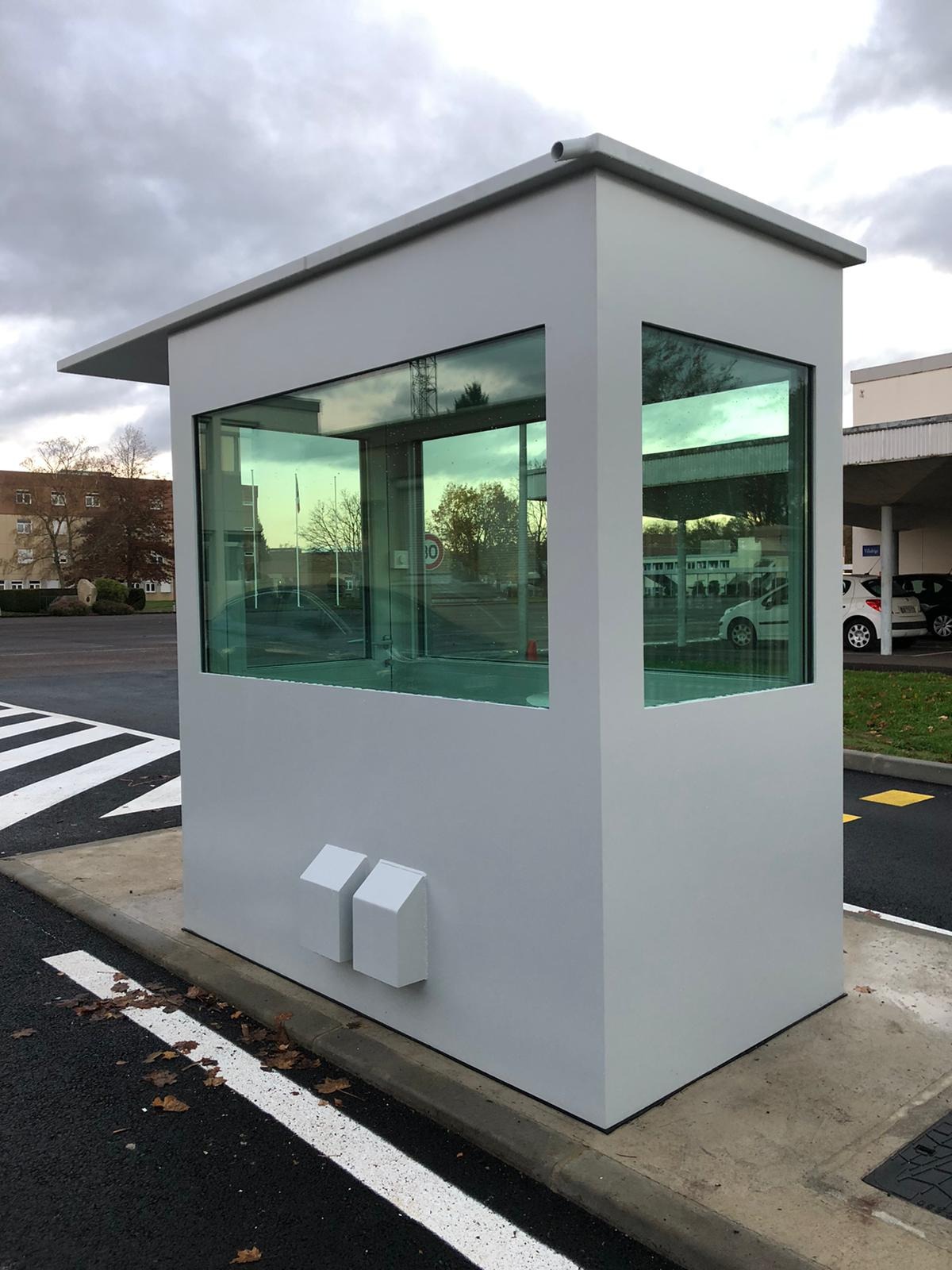 Nowoczesna, szara budka wartownicza z dużymi oknami, z widokiem na parking i budynki w tle, sfotografowana w pochmurny dzień.