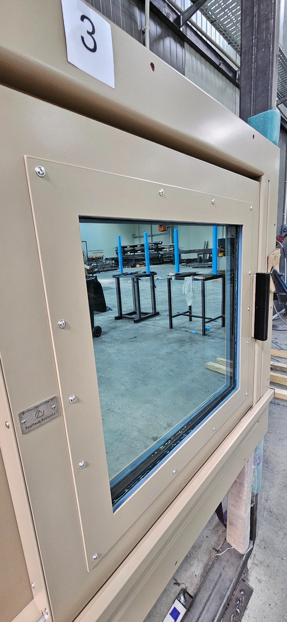 Beżowa, metalowa konstrukcja z szybą, zabezpieczona śrubami, z widocznym numerem '3' na górze i logotypem producenta na boku, stojąca w hali produkcyjnej z widocznymi elementami metalowymi w tle.
