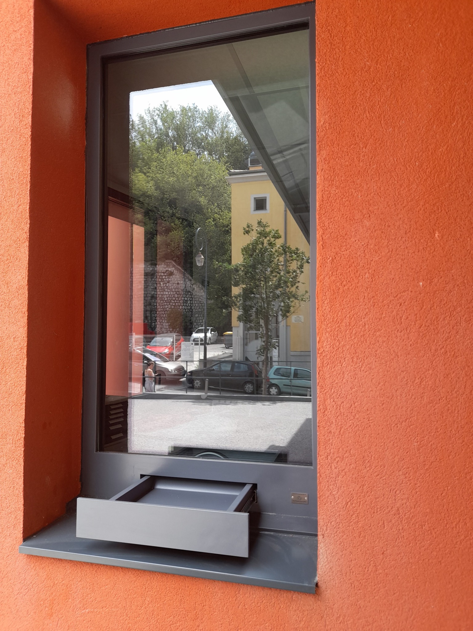 Okno z podajnikiem, szara rama, pomarańczowa ściana z fakturą, odbicie ulicy w szybie.