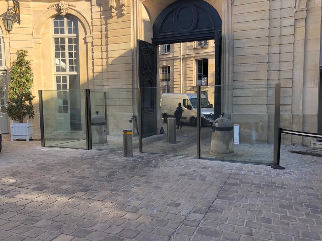 Szklana bariera z czarnymi ramami zamontowana przed wejściem do budynku o klasycznej architekturze, widoczny samochód dostawczy w tle.