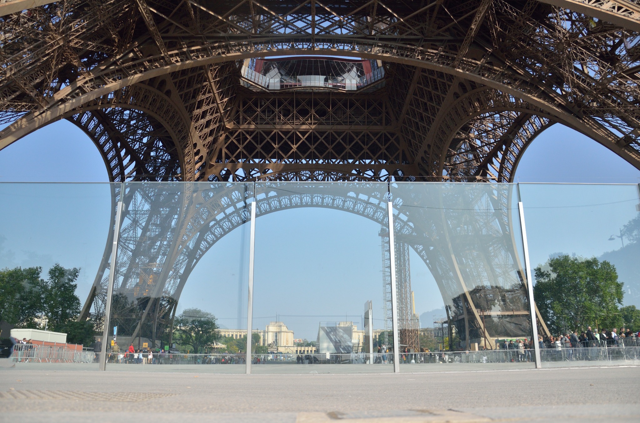 Widok na wieżę Eiffla przez szklaną barierę ochronną z metalowymi słupkami, z perspektywy niskiego kąta, ukazujący konstrukcję wieży i odbicie w szkle.