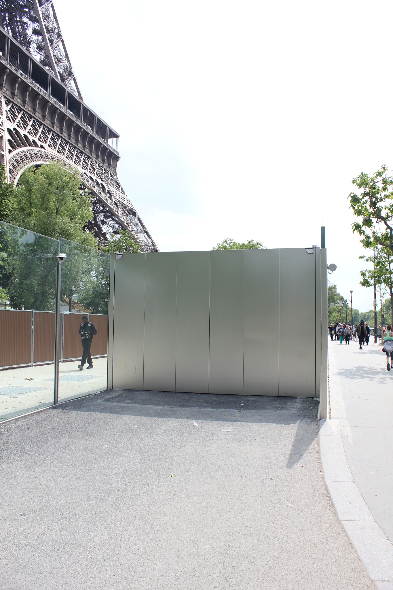 Nowoczesna, minimalistyczna, metalowa konstrukcja z przeszkleniami na tle wieży Eiffla i spacerujących ludzi.