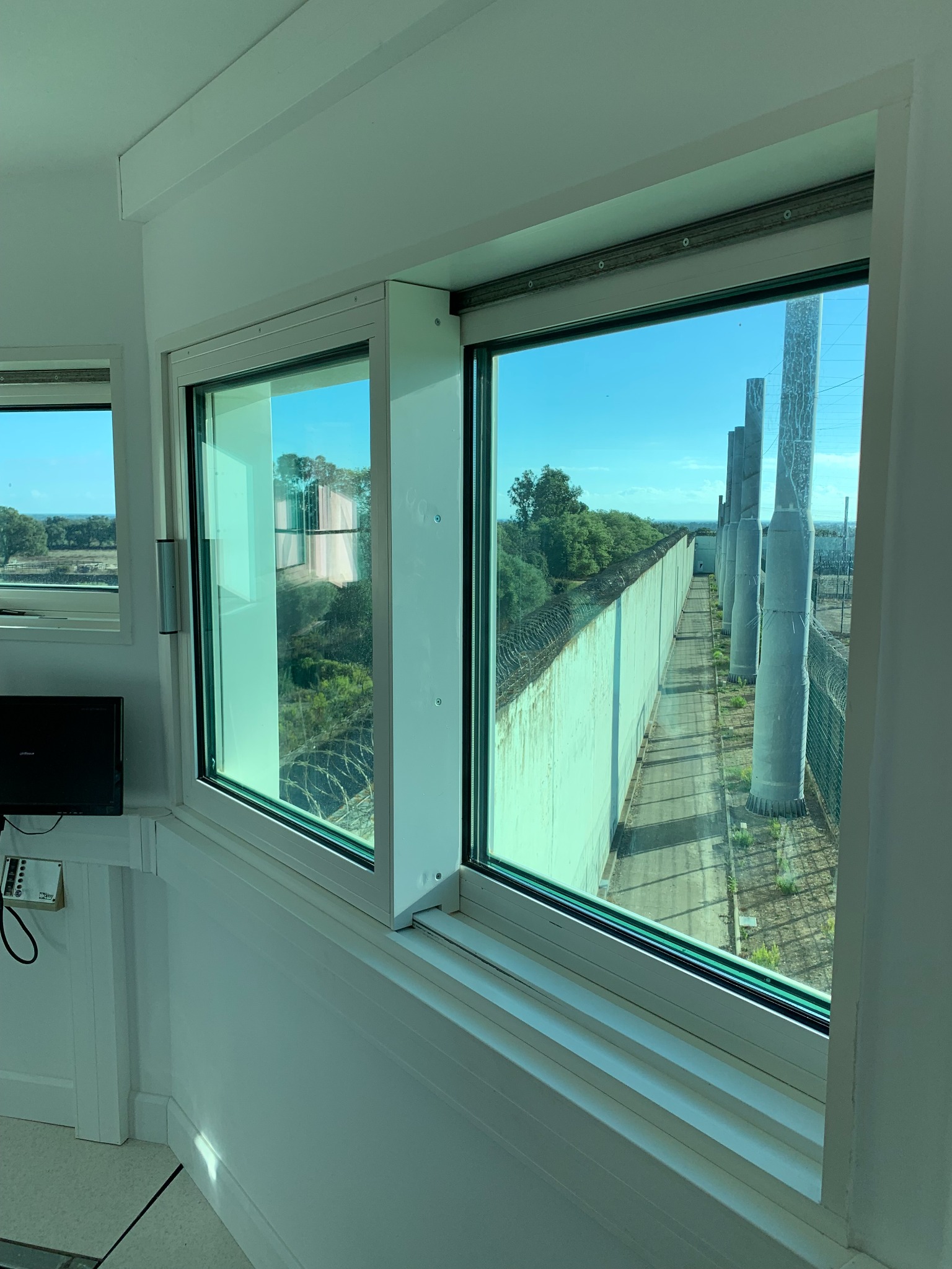 Widok z okna na mur z drutem kolczastym i betonowe słupy, prawdopodobnie ogrodzenie terenu chronionego.