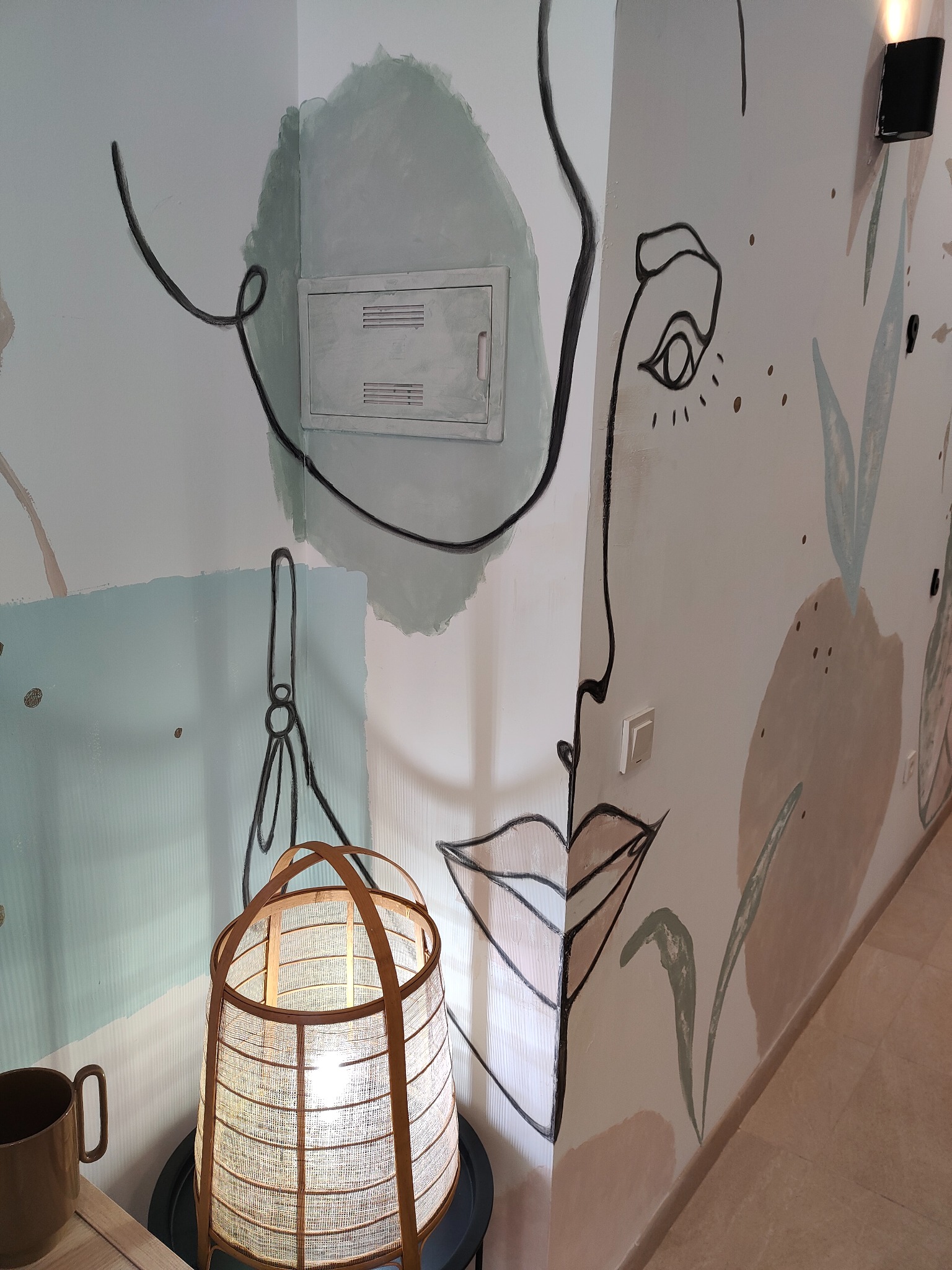 Ściana z abstrakcyjnym malunkiem przedstawiającym twarz, geometryczne kształty i roślinne motywy, oświetlona lampą w stylu boho, z widocznym fragmentem instalacji elektrycznej.