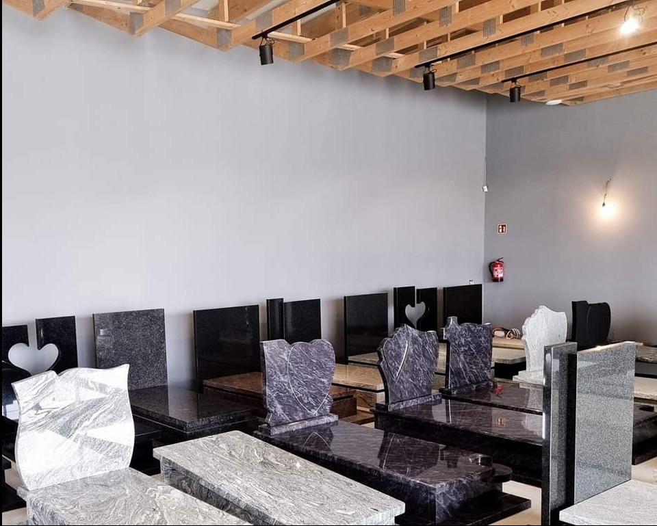 Wystawa różnorodnych, gotowych pomników z kamienia naturalnego, prezentowanych w zamkniętym showroomie z surowym, drewnianym stropem i szarą ścianą w tle.