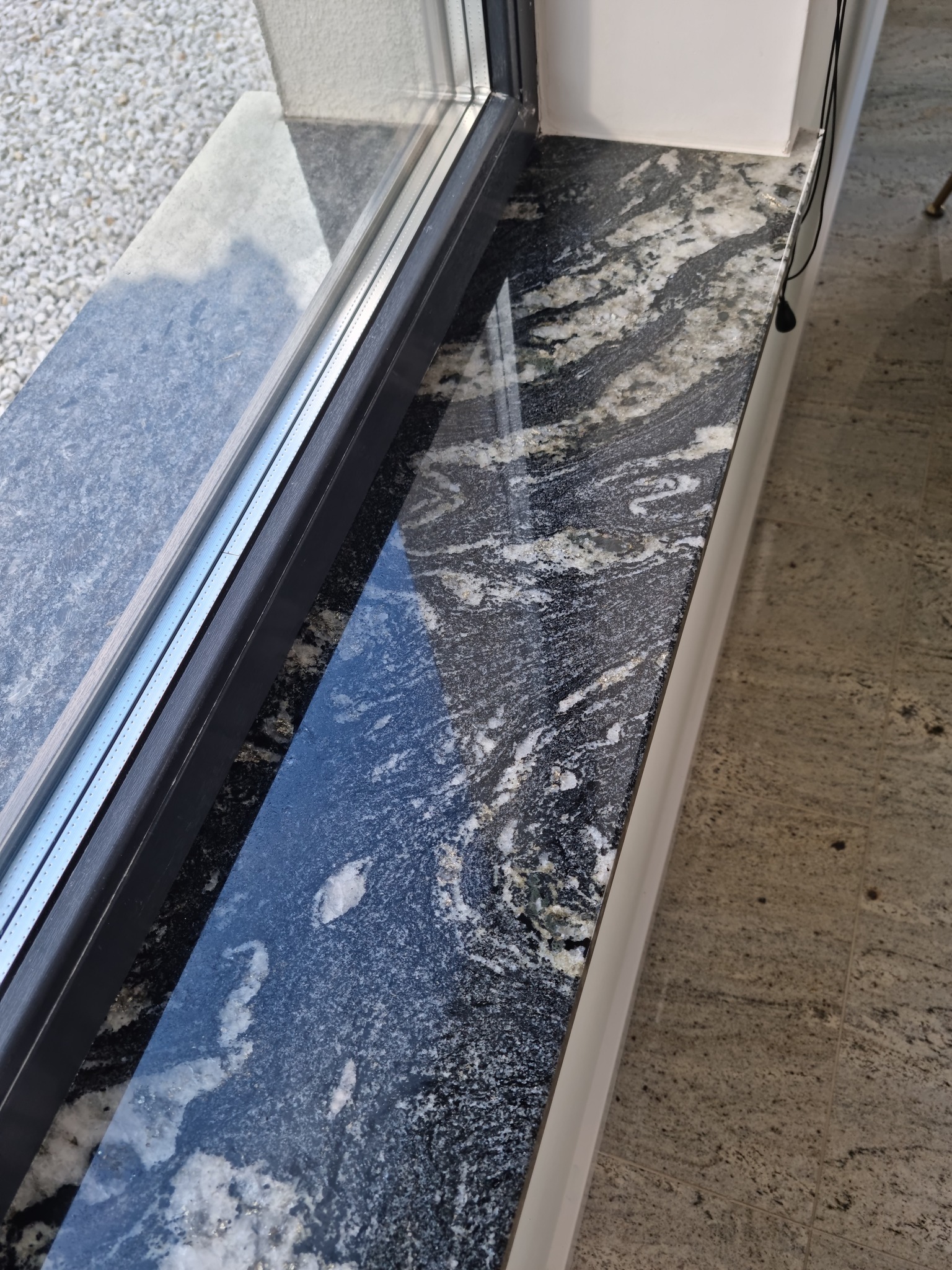 Granitowy parapet okienny o czarno-białej strukturze, widoczny w zbliżeniu, z refleksami światła na polerowanej powierzchni, obok okna z szarą ramą.