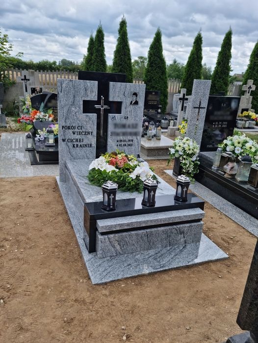Granitowy pomnik z czarnymi elementami, zdobiony wieńcem z jasnych kwiatów i lampionami, na tle cmentarza z tujami.