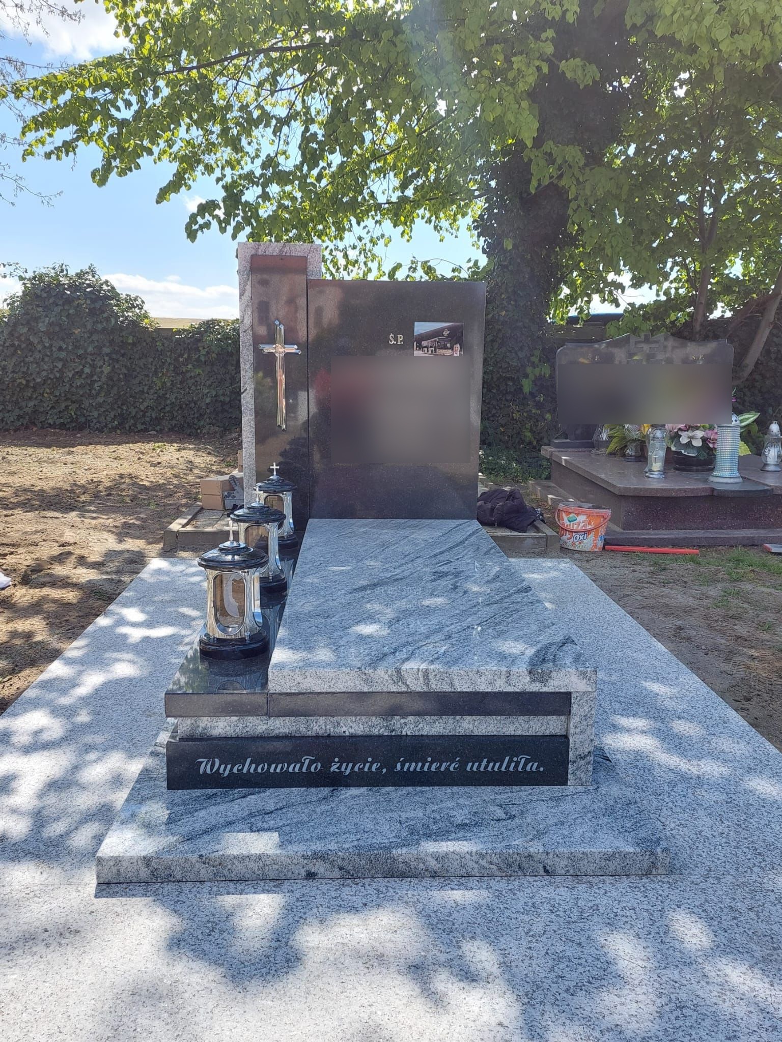 Polerowany, szary nagrobek z ciemnoszarą tablicą, srebrnym krzyżem i trzema lampionami, na cmentarzu w słoneczny dzień.