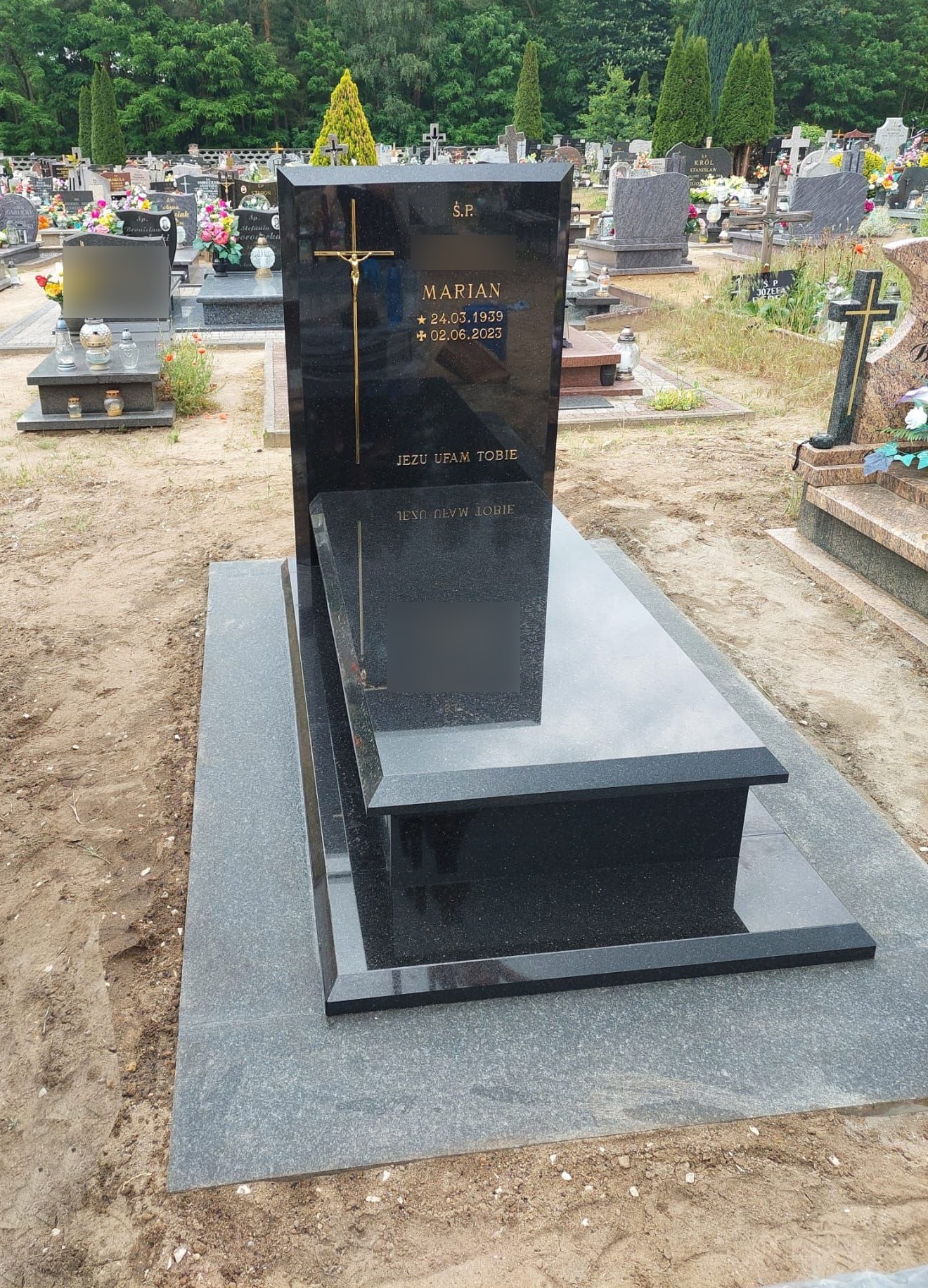 Czarny, polerowany pomnik z krzyżem i inskrypcją 'Marian 24.03.1939 + 02.06.2023' na tle cmentarza z innymi nagrobkami i drzewami.