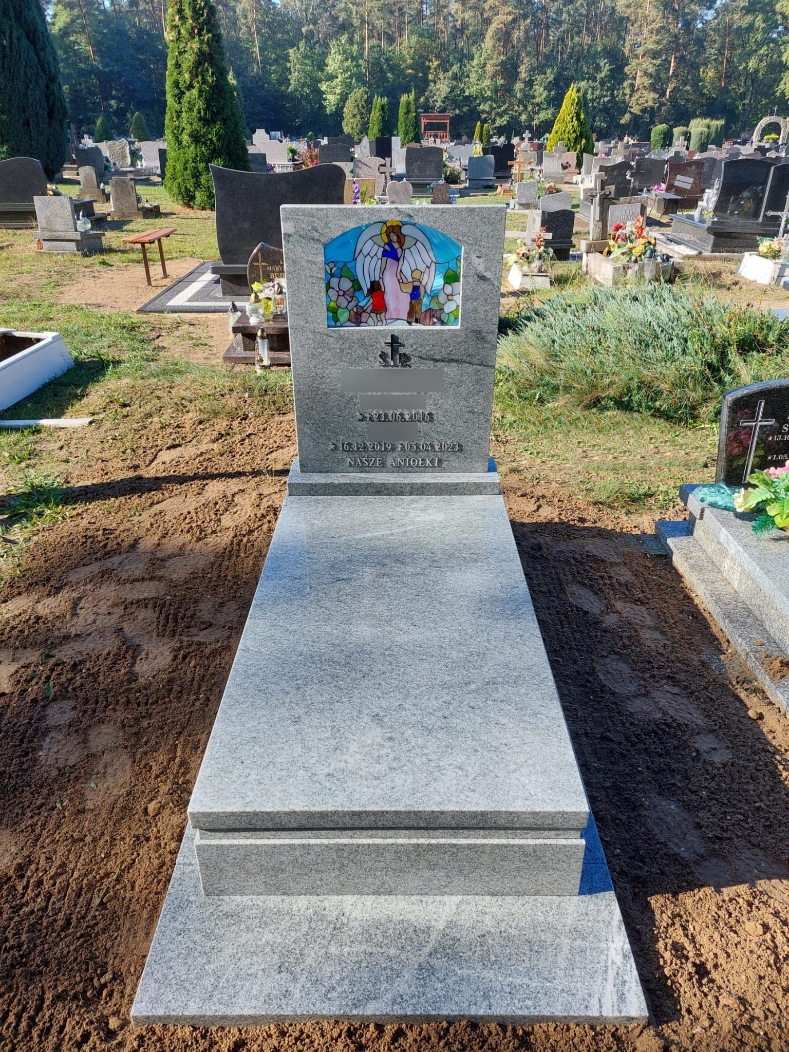Jasnoszary pomnik z kolorową mozaiką przedstawiającą anioła i dzieci, umieszczony na cmentarzu z licznymi nagrobkami w tle.