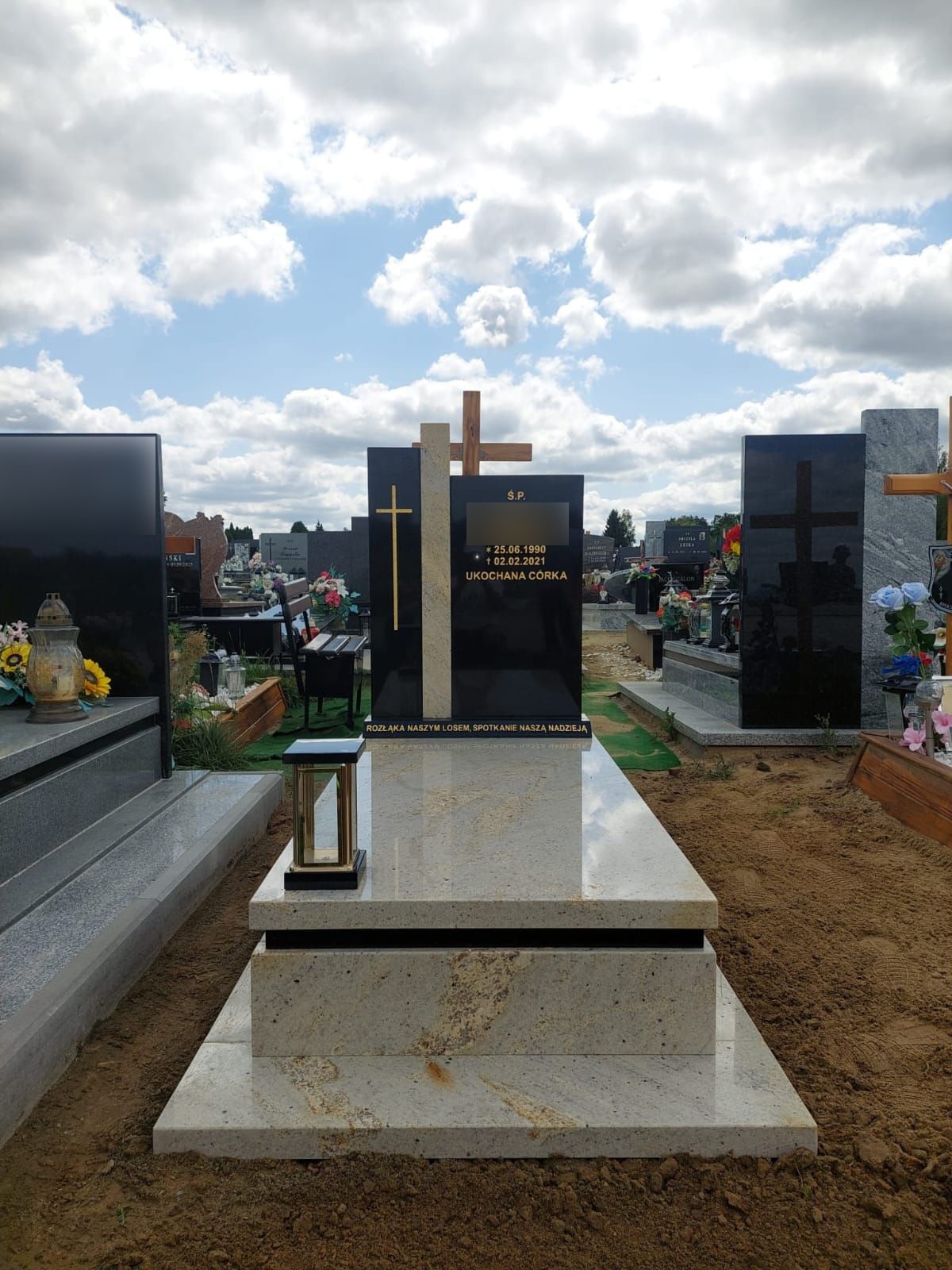 Jasny nagrobek z czarną tablicą i drewnianym krzyżem na tle cmentarza pod błękitnym niebem z chmurami.