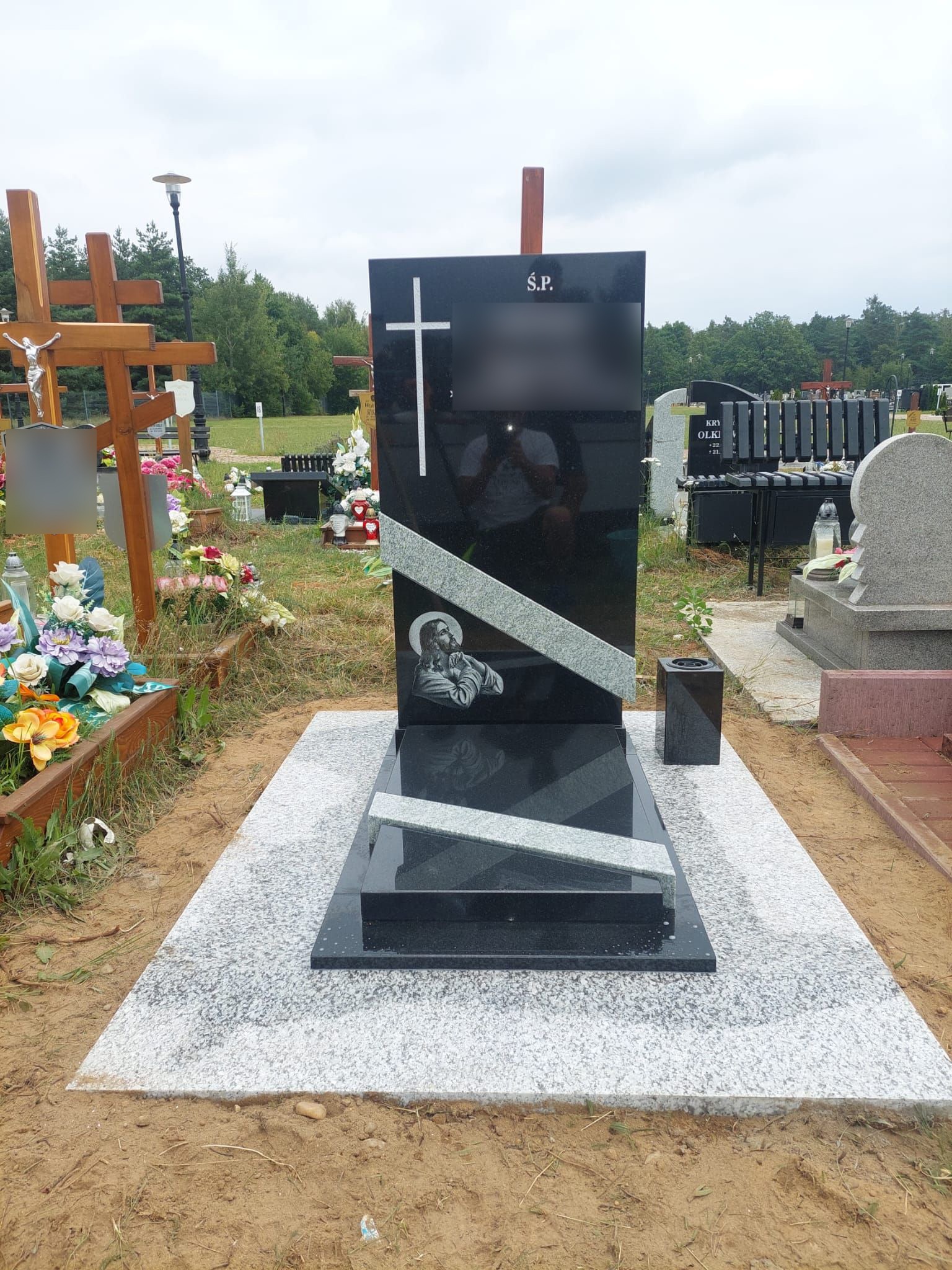 Czarny, polerowany pomnik z krzyżem i wizerunkiem modlącego się Jezusa, ukośny jasnoszary pas dekoracyjny, otoczenie cmentarza z widocznymi krzyżami i innymi nagrobkami.