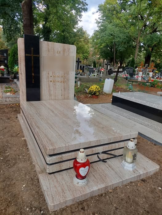 Jasny, warstwowy pomnik z czarnym akcentem krzyża i datami, ozdobiony dwoma lampionami w kolorze czerwonym i białym, na tle cmentarza z zielenią drzew.