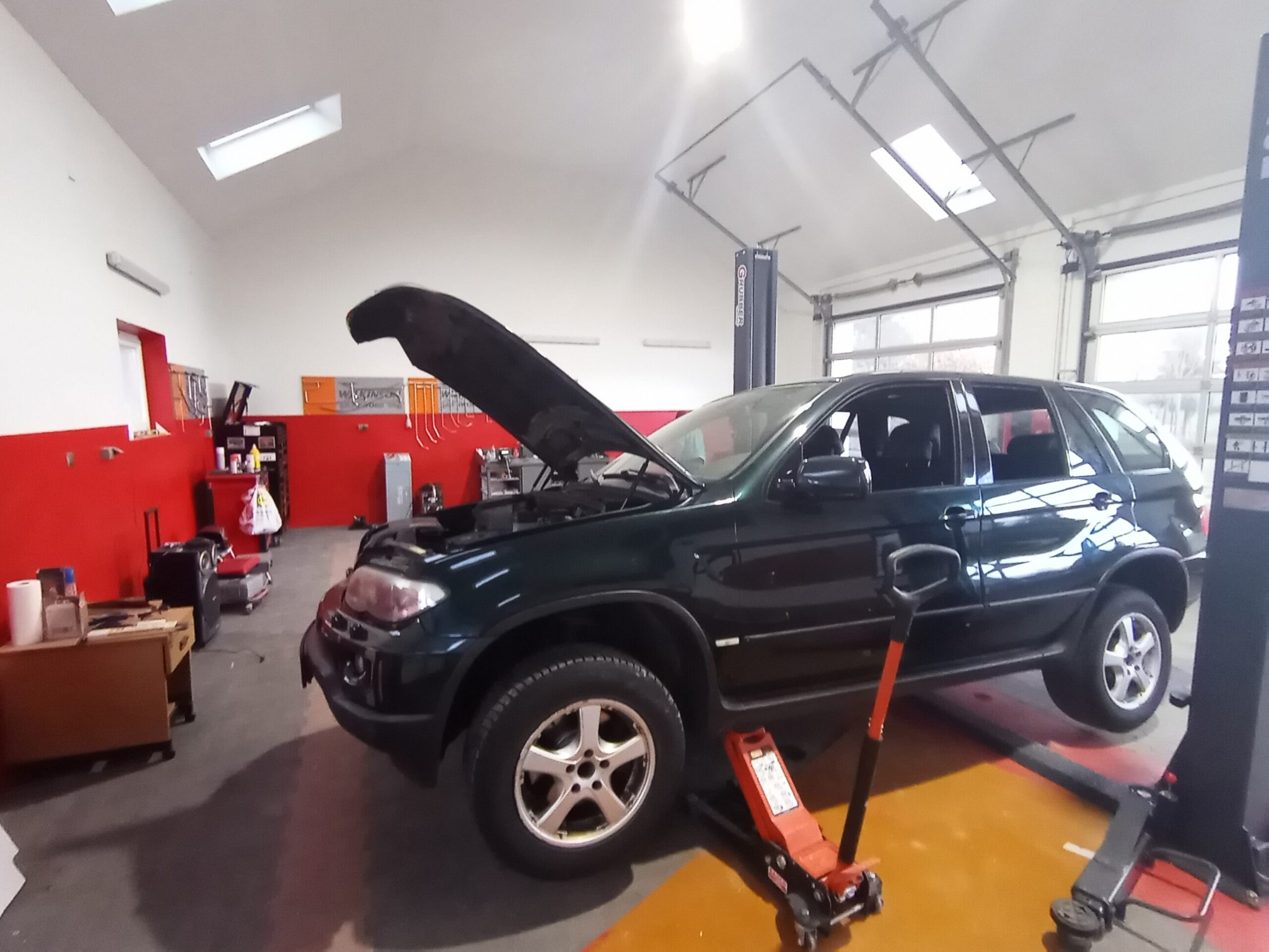 Ciemnozielony SUV na podnośniku w warsztacie, otwarty silnik, widoczny podnośnik hydrauliczny, jasne oświetlenie sufitowe.