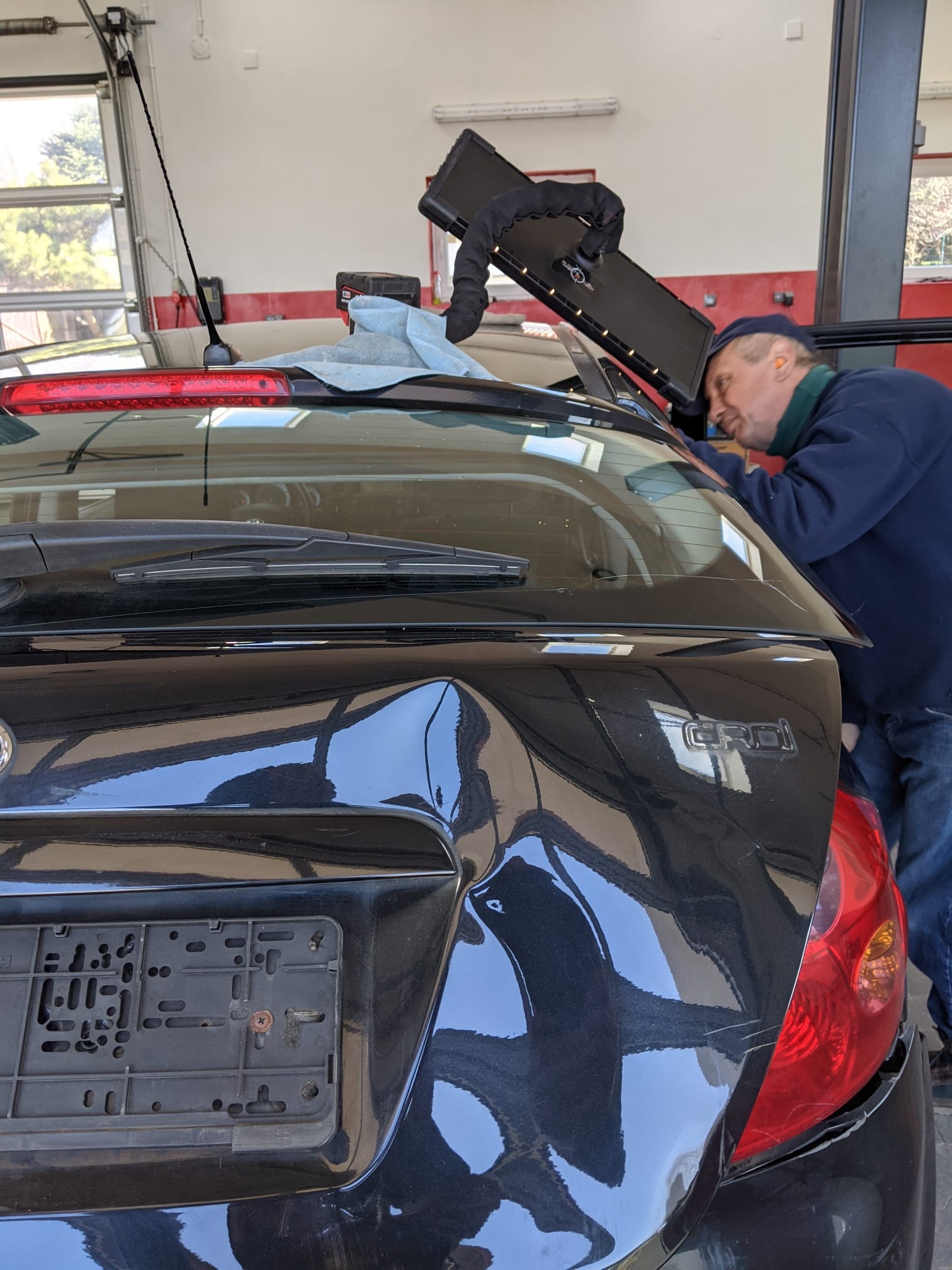 Czarny samochód z widocznym wgnieceniem na klapie bagażnika, technik w niebieskiej bluzie i czapce pracuje przy użyciu specjalistycznej lampy inspekcyjnej, widoczne narzędzia i elementy warsztatu...