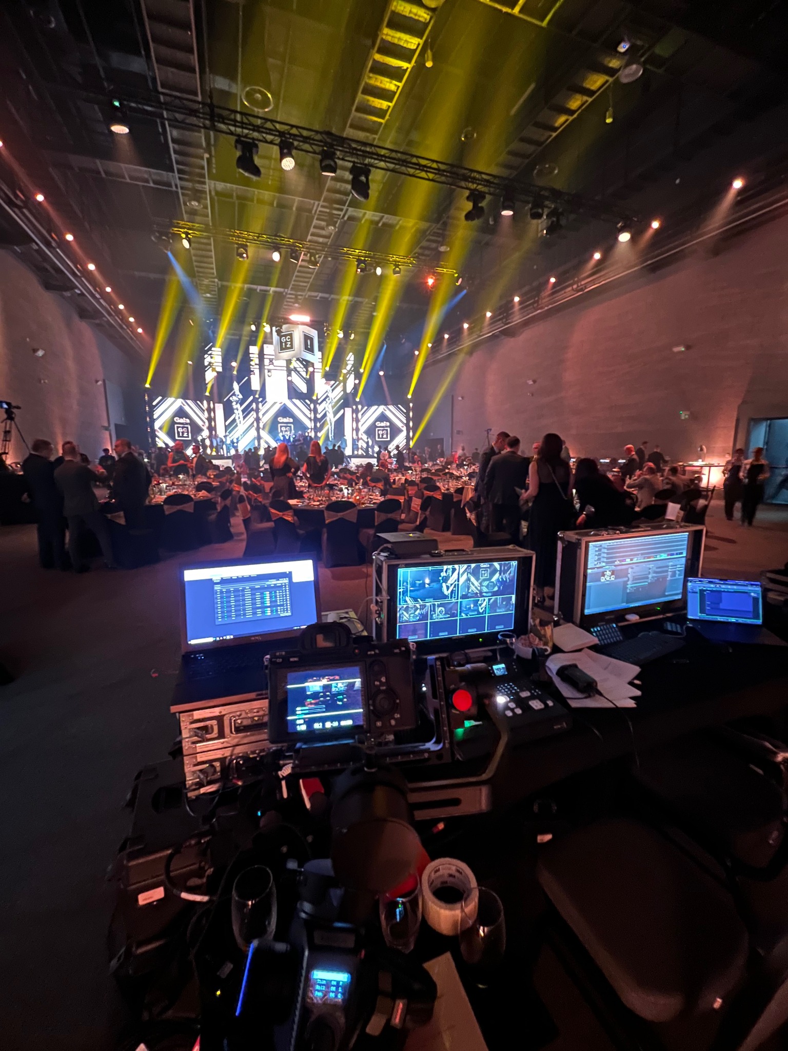 Sala bankietowa z oświetleniem scenicznym, widok zza stanowiska realizatora z monitorami i laptopem, tłum gości w tle. Żółte światła skierowane na scenę.
