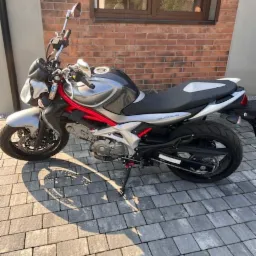 Szary motocykl Suzuki Gladius z czerwonymi elementami ramy zaparkowany na kostce brukowej w Zawierciu, na tle ceglanego budynku.