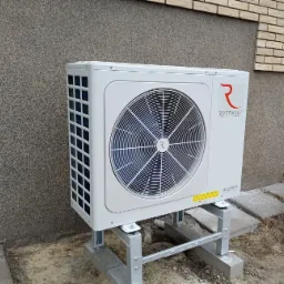 Biała pompa ciepła Rotenso Aquami zamontowana na metalowej konstrukcji przed ścianą budynku z szarą elewacją i fragmentem ściany z cegły.