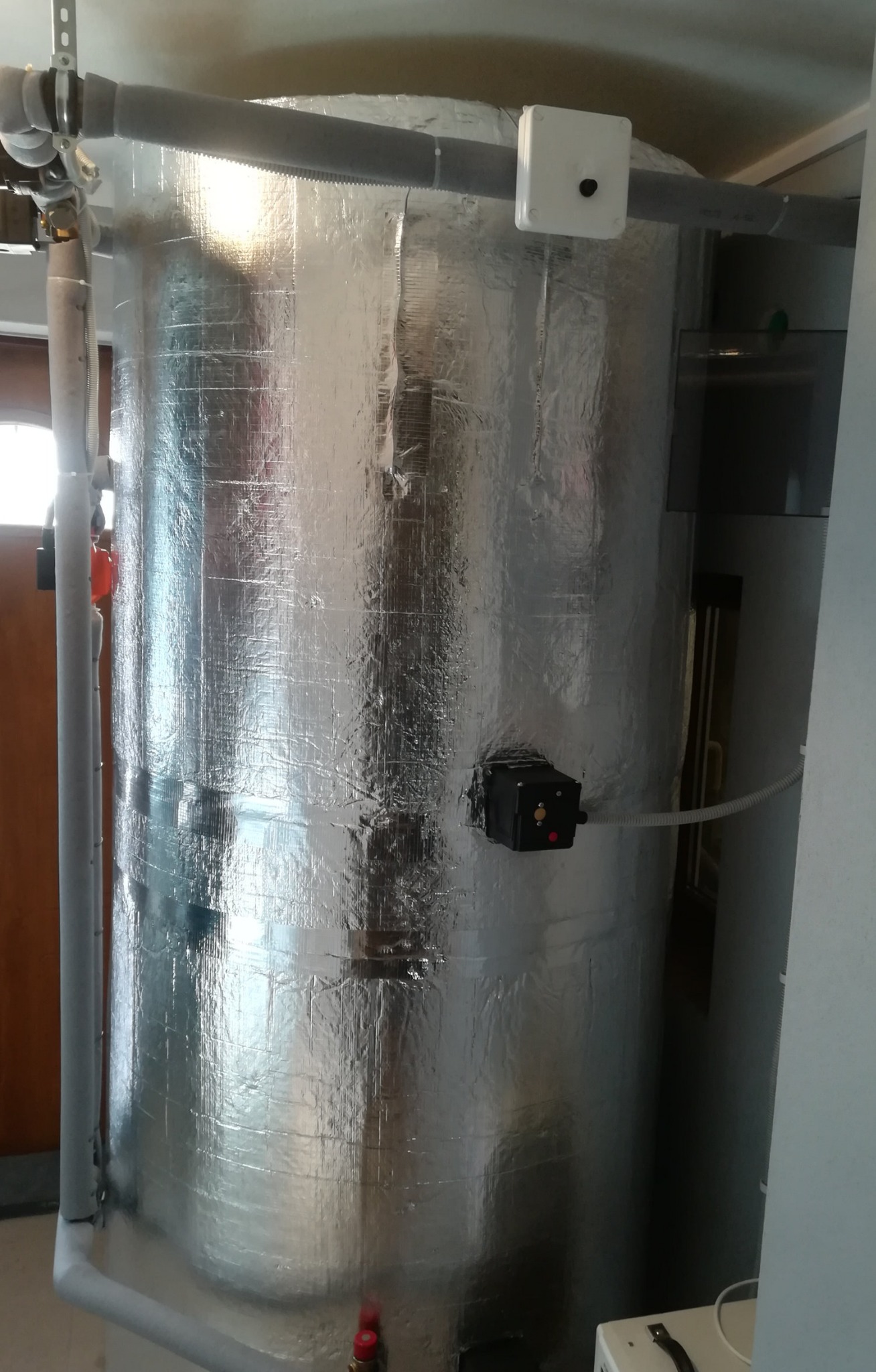 Izolowany srebrną folią zbiornik akumulacyjny z podłączonymi rurami i skrzynką sterowniczą, widoczne elementy instalacji w pomieszczeniu.