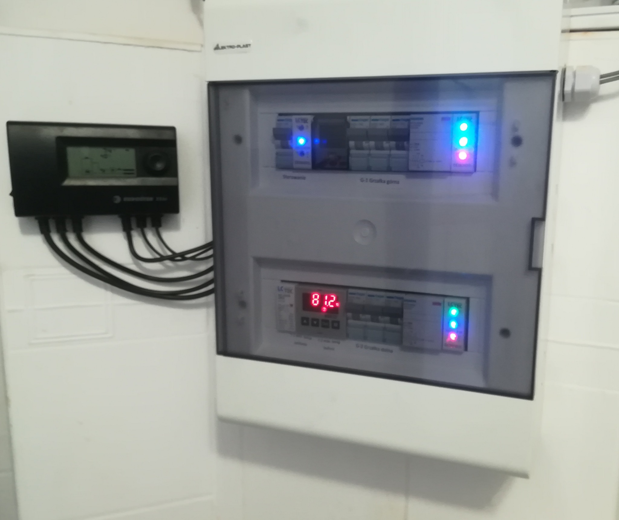 Rozdzielnica elektryczna z bezpiecznikami i wyświetlaczem LED wskazującym parametry pracy instalacji, zamontowana na białej ścianie.