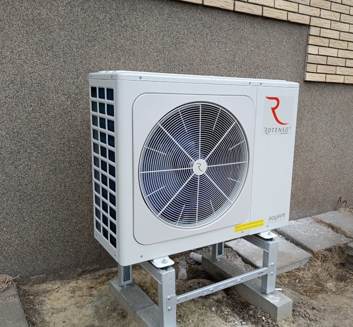 Biała pompa ciepła Rotenso Aquami zamontowana na metalowej konstrukcji przed ścianą budynku z szarą elewacją i fragmentem ściany z cegły.