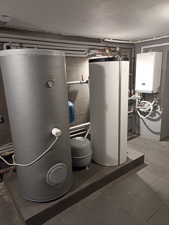 Szara i biała instalacja grzewcza w piwnicy z widocznymi rurami na ścianie i suficie, mały niebieski zbiornik wyrównawczy obok dużego szarego zbiornika.