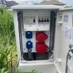 Otwarta skrzynka z bezpiecznikami i gniazdami siłowymi (czerwone i niebieskie) na zewnątrz, otoczona zielenią. Widoczne elementy instalacji elektrycznej.