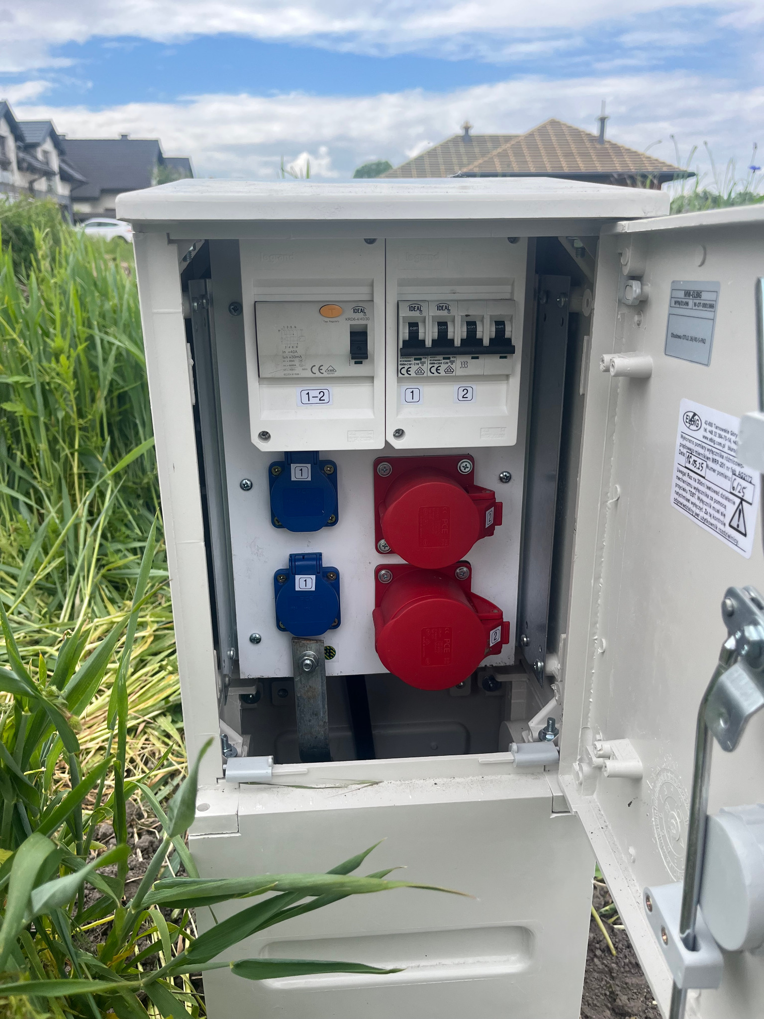 Otwarta skrzynka z bezpiecznikami i gniazdami siłowymi (czerwone i niebieskie) na zewnątrz, otoczona zielenią. Widoczne elementy instalacji elektrycznej.