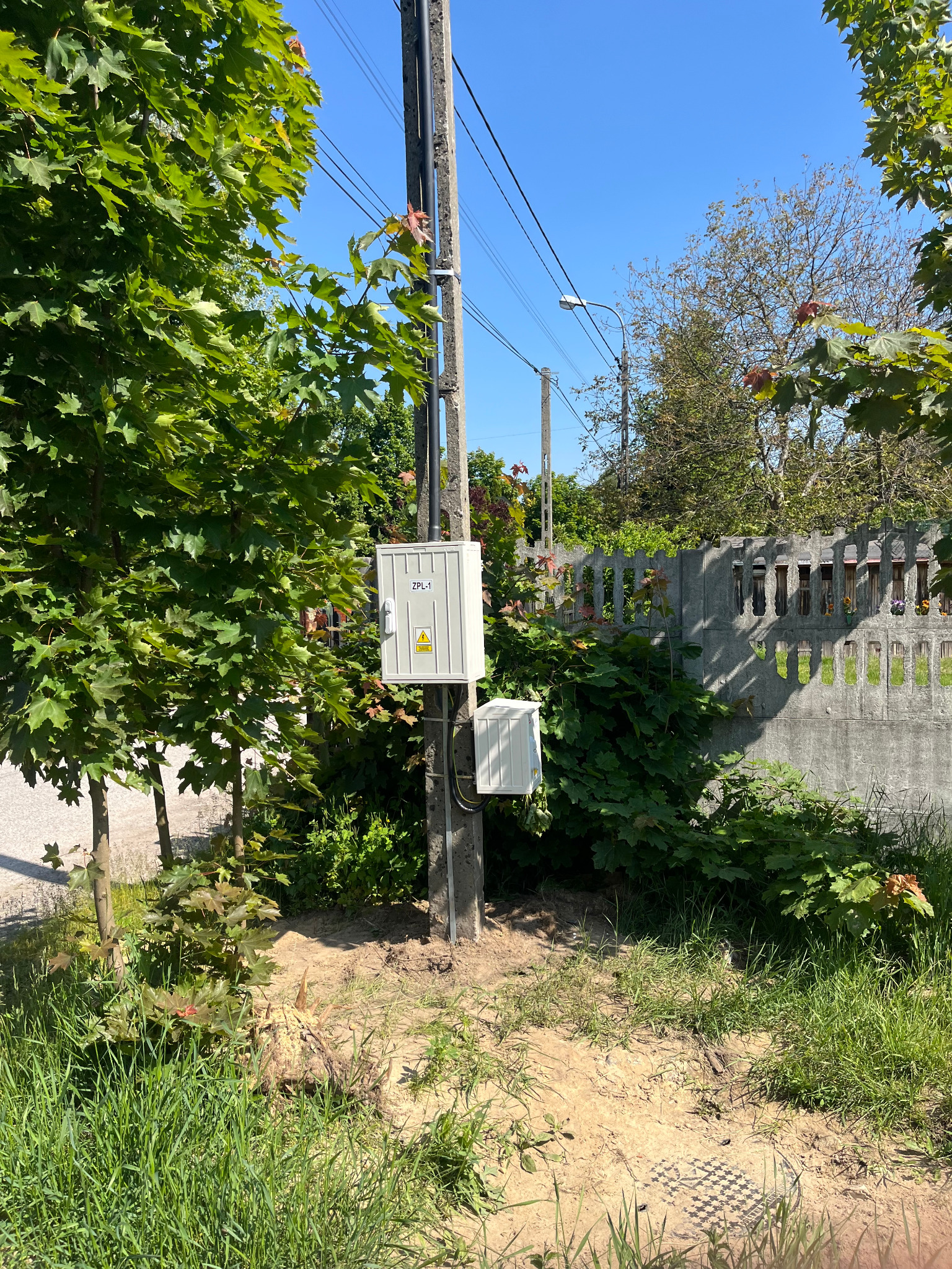 Słup energetyczny z dwiema skrzynkami ZPL-1 na tle zieleni i betonowego ogrodzenia, oświetlony jasnym słońcem.