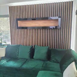 Szmaragdowa sofa narożna z weluru, z poduszkami, na tle ściany z drewnianych listew z czarną ramą i półką z drewna.