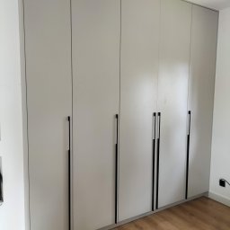 As Meble Radom - Jasnoszara szafa wnękowa z długimi, czarnymi uchwytami na tle drewnianej podłogi. Minimalistyczny design, proste linie, subtelne oświetlenie. Widok z dołu, lekko z prawej strony.