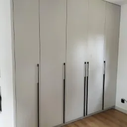 Jasnoszara szafa wnękowa z długimi, czarnymi uchwytami na tle drewnianej podłogi. Minimalistyczny design, proste linie, subtelne oświetlenie. Widok z dołu, lekko z prawej strony.