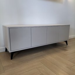 As Meble Radom - Minimalistyczna szafka RTV z czterema drzwiczkami w odcieniach szarości, na niskich, czarnych nóżkach, na tle białej ściany i parkietu w jodełkę.