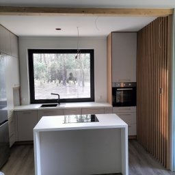 As Meble Radom - Jasna kuchnia z wyspą i dużym oknem z widokiem na las, drewniana ściana z listew, płyta indukcyjna na wyspie.